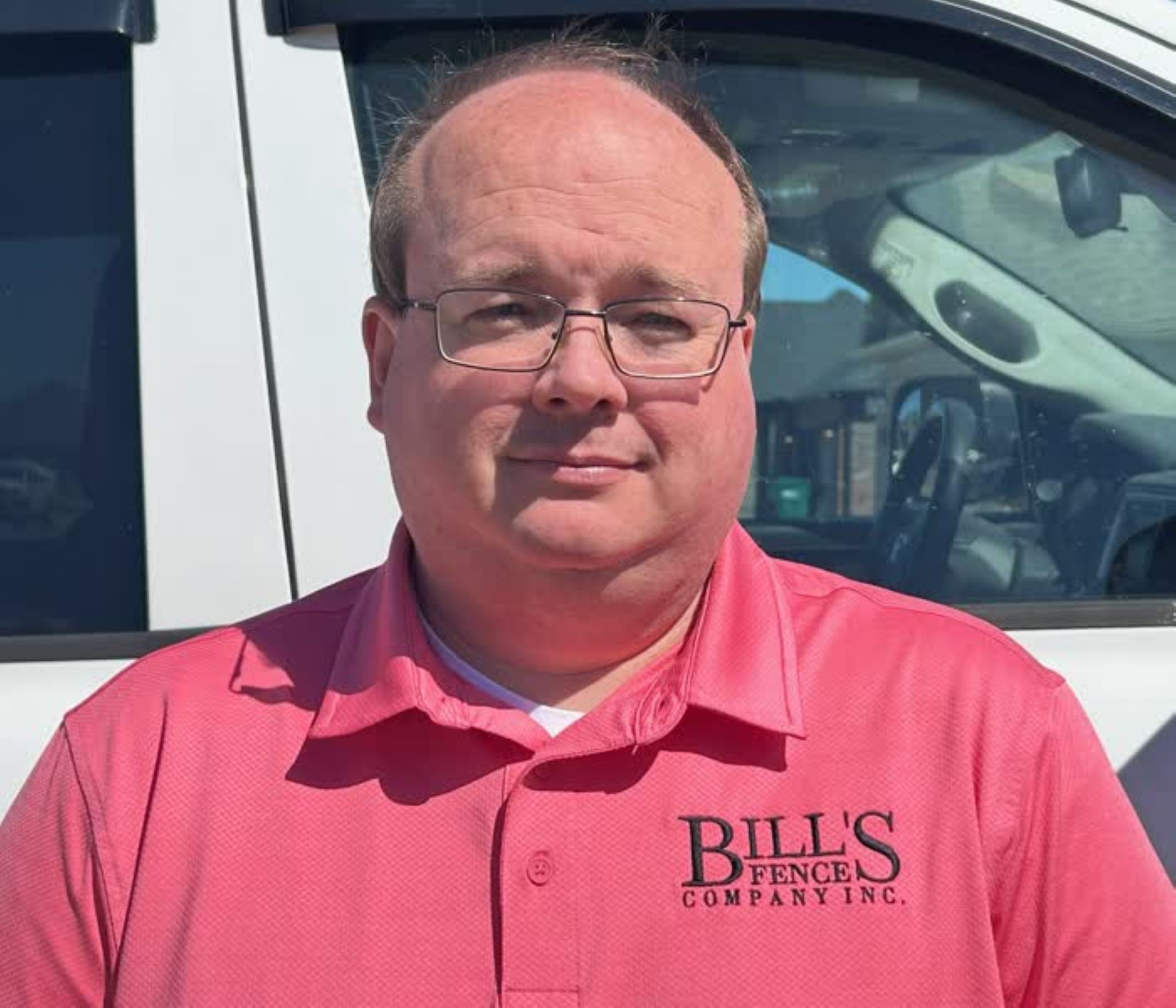 A man wearing glasses and a pink “BILL'S FENCE CO.” polo shirt stands in front of a white vehicle on a sunny day.