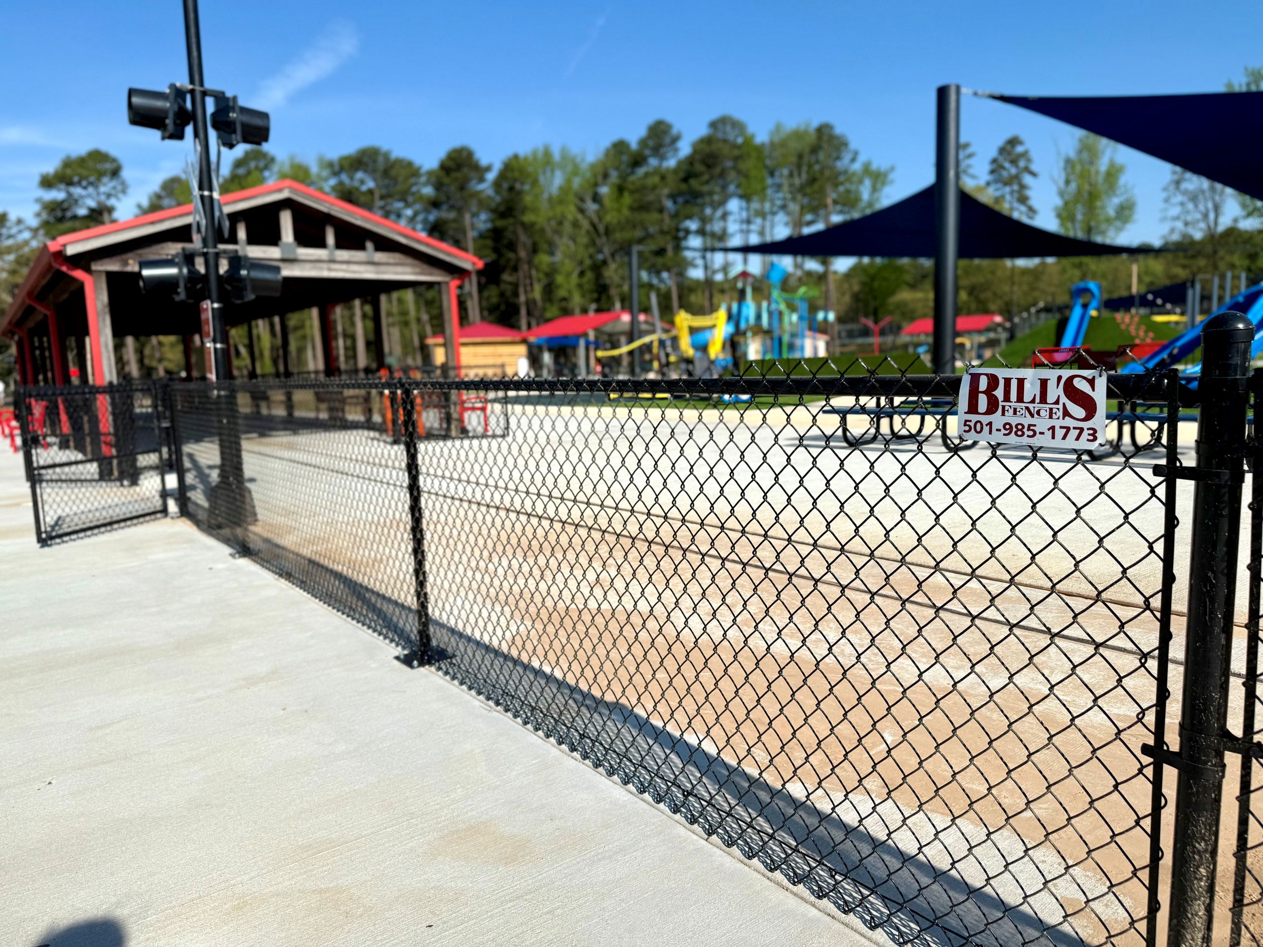 A chain-link fence surrounds an outdoor playground with colorful equipment and shaded picnic areas. A sign on the fence reads Bills Fence with a phone number. Trees, a pavilion, and quality chain link fencing are visible under a clear sky.