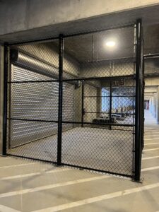 A chain-link fence with a gate blocks off part of an industrial corridor with concrete walls and ceiling, overhead lights, and a metal garage door in the background.