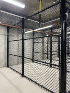 A fenced storage area with black chain-link panels and a concrete floor, enclosed within an industrial building with exposed pipes and ceiling fixtures.