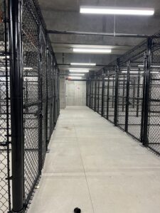 A concrete hallway with overhead lights, lined on both sides by tall black chain-link fencing, leading to a closed gray door at the end. The scene appears industrial and secure.