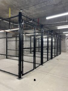 A large indoor area with concrete floors and ceiling, featuring a black chain-link fenced enclosure with multiple sections under bright fluorescent lighting. Industrial pipes and wiring are visible above.