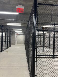 A hallway with concrete floors and walls, lined with black chain-link storage cages. Overhead fluorescent lights brighten the space, and a red EXIT sign hangs from the ceiling near the end of the hall.