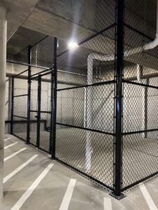 A black chain-link fence forms an enclosed area in a concrete parking garage, with white painted lines on the floor and exposed pipes along the walls and ceiling. Bright overhead lighting casts shadows.