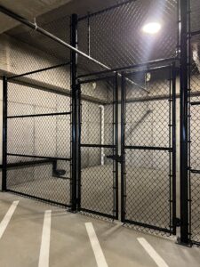A tall black chain-link fence with a gate is installed inside a concrete parking garage, separating an area from the rest of the space. Overhead lighting illuminates the scene. White parking lines are visible on the floor.