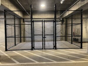 A locked black chain-link fence enclosure stands in a parking garage with concrete walls, a ceiling light above, and white diagonal lines painted on the ground.