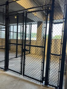 A metal chain-link fence with a locked gate encloses a concrete area in a building, lit by overhead lights and natural light coming from outside.