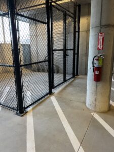 A parking garage with a fire extinguisher mounted on a concrete pillar. A black chain-link fence encloses a storage area in the background. White parking lines are painted on the concrete floor.