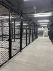 A clean, empty indoor space with tall, black chain-link cage enclosures along the left side and bright overhead lighting. There is a concrete floor and an elevator at the end of the hallway.