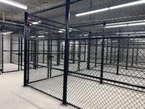 A large, empty indoor space with multiple black chain-link fenced enclosures separated by metal posts, illuminated by bright overhead fluorescent lights. The floor is bare concrete.