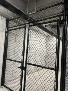 A black chain-link fence gate inside a concrete room, with overhead pipes and exposed ceiling. The area appears industrial or utilitarian.
