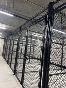A row of tall black metal chain-link cages with locked gates inside a concrete room, lit by overhead fluorescent lights.