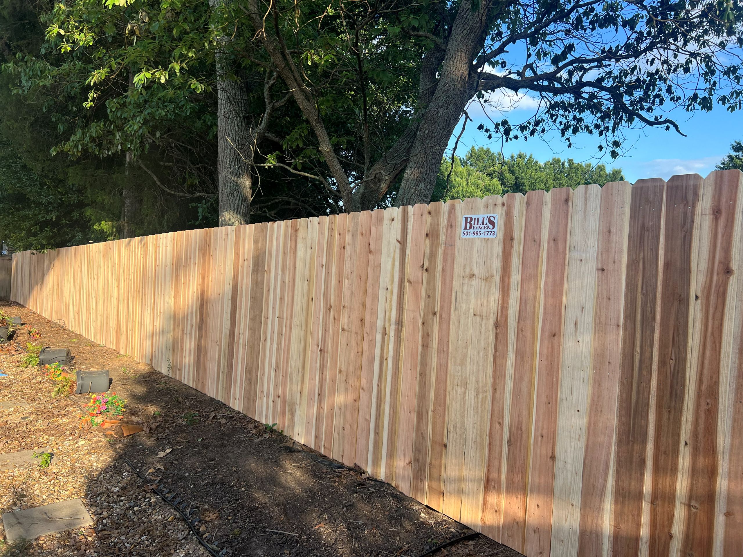 A tall, wooden fence runs alongside a garden bed with trees and greenery in the background. A small sign reading Bills Fence Company is attached to the fence, standing out beneath a partly cloudy sky.