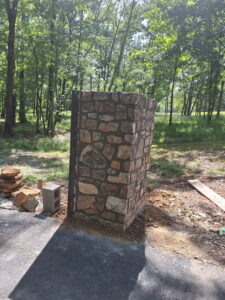 A small stone structure with irregular stones stands on a paved area near a grassy, wooded area in sunlight. Nearby are a cinder block, loose stones, and wooden planks.