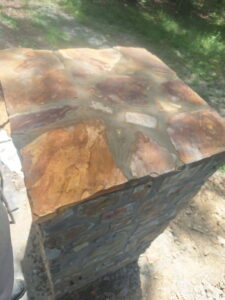 A stone pillar with a flat top made of various colored stones and visible mortar, standing outdoors on rocky ground.