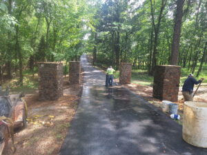 Two workers are building a stone entryway with four pillars at the end of a newly paved driveway surrounded by trees in a wooded area.