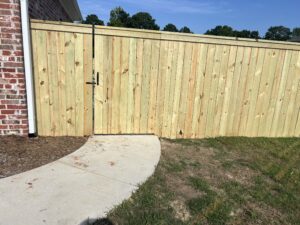 A new wooden fence with a closed gate stands next to a brick house, beside a curved concrete walkway bordered by grass and mulch.