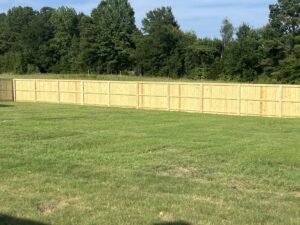 A long, light wooden fence stands on green grass with dense trees and bushes in the background under a blue sky.