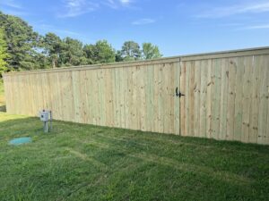 A tall wooden privacy fence with a gate and a metal latch stands on green grass, with trees and blue sky in the background. An electrical box and a green circular cover are visible in the lawn.