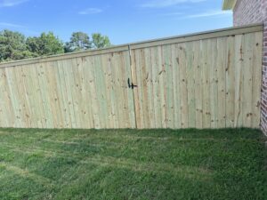 A tall wooden fence with a closed gate and black handle stands next to a brick building, enclosing a grassy yard on a sunny day. Trees and blue sky are visible in the background.
