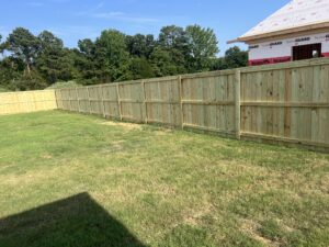 A newly built wooden privacy fence runs along the edge of a green grassy backyard, with trees in the background and a partially constructed house visible on the right.