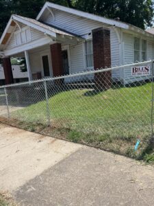 A white house with a brick porch sits behind a chain-link fence. A BILLS sign with a phone number is attached to the fence. The yard is grassy and a paved sidewalk is in front.