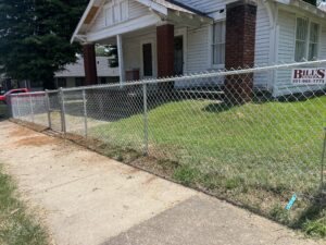 A chain-link fence borders a grassy yard in front of a white house with red brick columns. A sign on the fence reads Bills Fence with a phone number. A sidewalk runs alongside the fence.