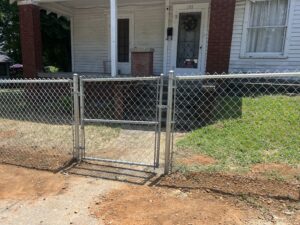 A chain-link fence with a closed gate blocks a sidewalk leading to the porch of a white house with brick columns and the house number 201. Grass and dirt surround the walkway outside the fence.