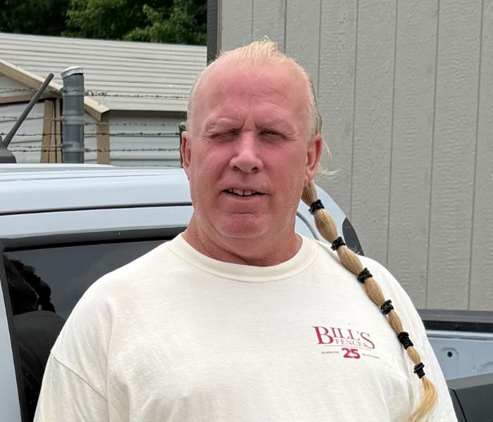 A man with a long blond braid and receding hairline stands outside near a gray building, wearing a white Bills Fence T-shirt.
