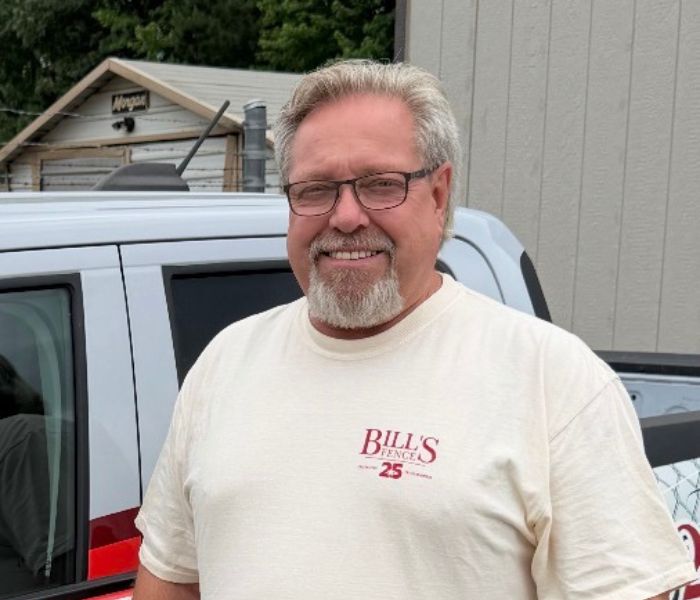 A smiling man with glasses and a beard wears a BILLS FENCE 25 t-shirt, standing outside next to a gray vehicle and a building.