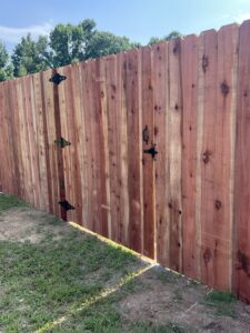 A tall wooden fence with a closed gate, black metal hinges, and latch. Sunlight shines through the gap beneath the gate. Green grass and trees are visible in the background.
