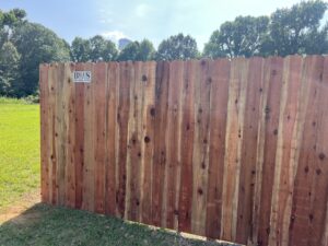 A tall wooden fence stands on grass with a small sign reading Bills 615-555-1733 in the upper left corner. Trees and a blue sky are visible in the background.