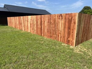 A new wooden privacy fence surrounds a grassy yard under a blue sky, with a dark-roofed building visible behind the fence.