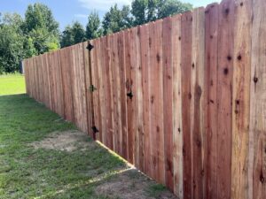 A tall wooden privacy fence with vertical planks and visible hinges stands on grass, with trees and greenery in the background under a clear sky.