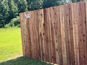 A wooden privacy fence stands on grass with a sign reading “Buss Fence Co.” Sunlight and trees are visible in the background.