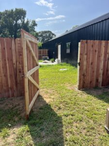 A wooden gate stands open, leading to a grassy yard with a black building and scattered white bags or objects under a sunny sky with trees in the background.