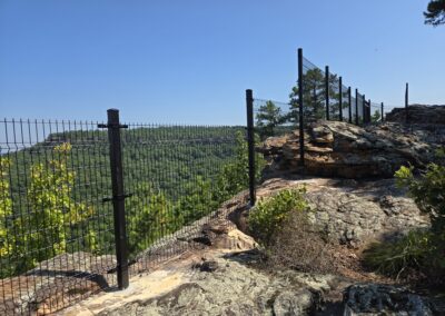 A metal fence runs along the edge of a rocky cliff, overlooking a lush green forest and distant hills under a clear blue sky.