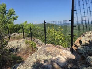 A black metal fence runs along the edge of a rocky cliff, overlooking a green forest and distant hills under a clear blue sky.