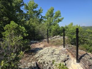 A rocky hilltop with black metal fencing and dense green trees under a clear blue sky. The fence runs along the edge, providing safety near the drop-off.