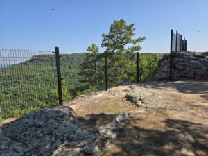 Rocky overlook with a metal fence, trees and green hills in the background under a clear blue sky. A couple of small birds are flying in the distance.