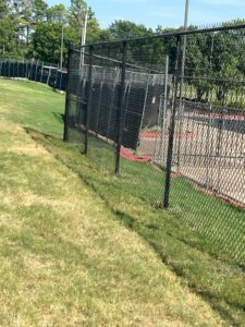 A black chain-link fence runs along the edge of a grassy field, separating it from a paved area and black barriers. Trees and light poles are visible in the background under a clear, sunny sky.