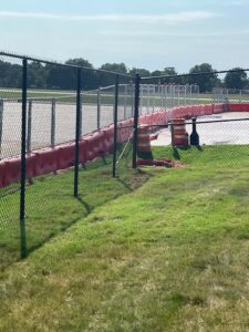 A chain-link fence lines a grassy area next to a road with red barriers and orange traffic barrels. Trees are visible in the background under a sunny sky.