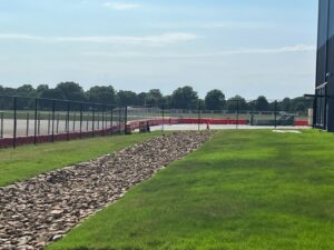 A race track with a gravel runoff area, green grass, and a fence alongside it. Trees are visible in the distance under a clear blue sky.