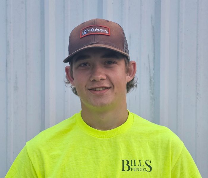 A young man wearing a brown Kubota cap and a neon yellow Bills Fence T-shirt stands in front of a light-colored corrugated metal wall, smiling at the camera.