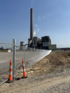 A power plant with a tall smokestack is seen behind a chain-link fence, with two orange traffic cones in front and clear blue sky above. Dirt and construction areas are in the foreground.