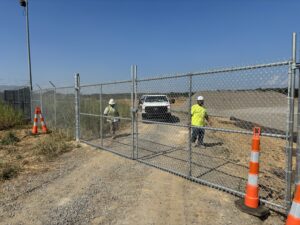 Two construction workers in safety vests and helmets stand near a chain-link gate. An orange-vested worker kneels by the gate, while a white pickup truck and orange cones are visible in the background on a dirt road.