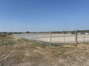A chain-link fence surrounds a large, cleared, gravel lot with construction vehicles in the distance under a clear blue sky. One orange traffic cone is near the fence corner. Sparse grass covers the foreground.