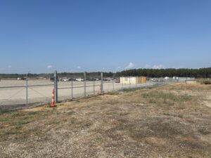 A fenced construction site with scattered vehicles, a few yellow containers, traffic cones, and sparse grass under a clear blue sky. Trees are visible in the background.