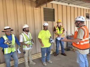 Five construction workers, wearing safety gear and holding items, stand by a metal building as one worker in an orange vest addresses the group.