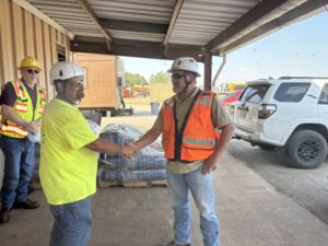 Two construction workers wearing safety vests and helmets shake hands outdoors, while another worker stands nearby holding papers. There is a white SUV and pallets of materials in the background.